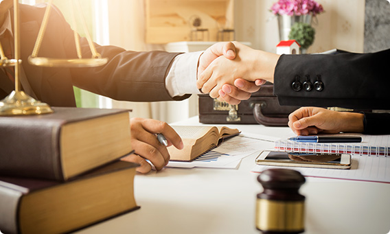 A lawyer and client shake hands over a desk cluttered with books, papers, and a gavel.